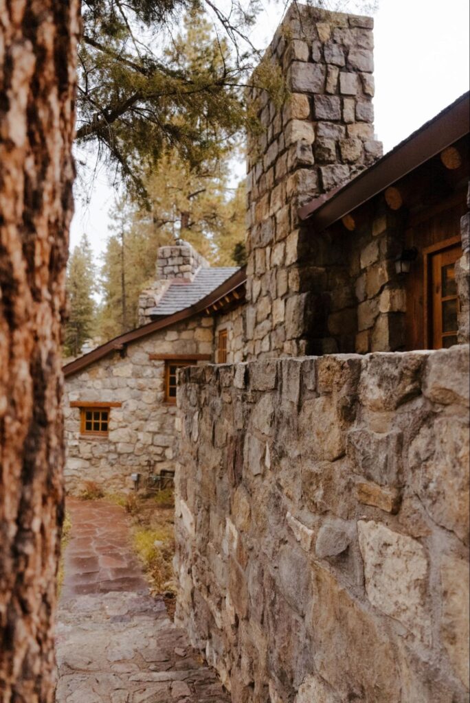 gorgeous outdoor destination wedding venue in santa fe that features a rustic stone building with wooden window frames and a chimney, set among tall pine trees. A stone walkway leads toward the building.