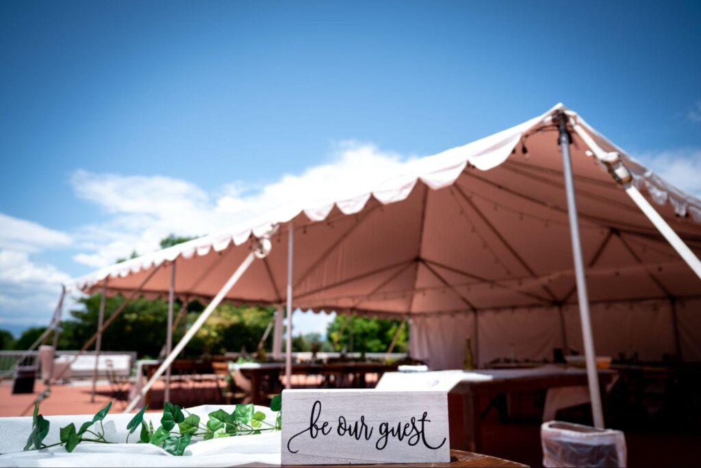 a large outdoor tent under a clear blue sky with a wooden sign in the foreground that reads "be our guest," accompanied by greenery as decoration.