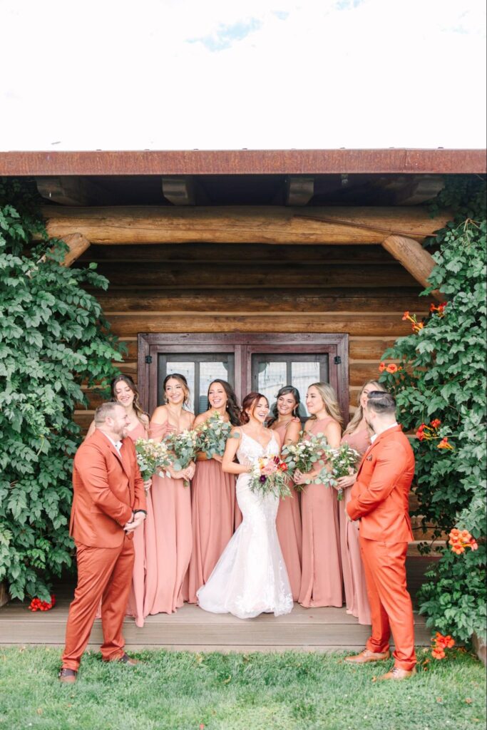 a smiling bride surrounded by her bridesmaids in matching blush dresses and bridesmen in rust-colored suits, standing in front of a rustic wooden cabin with lush greenery.