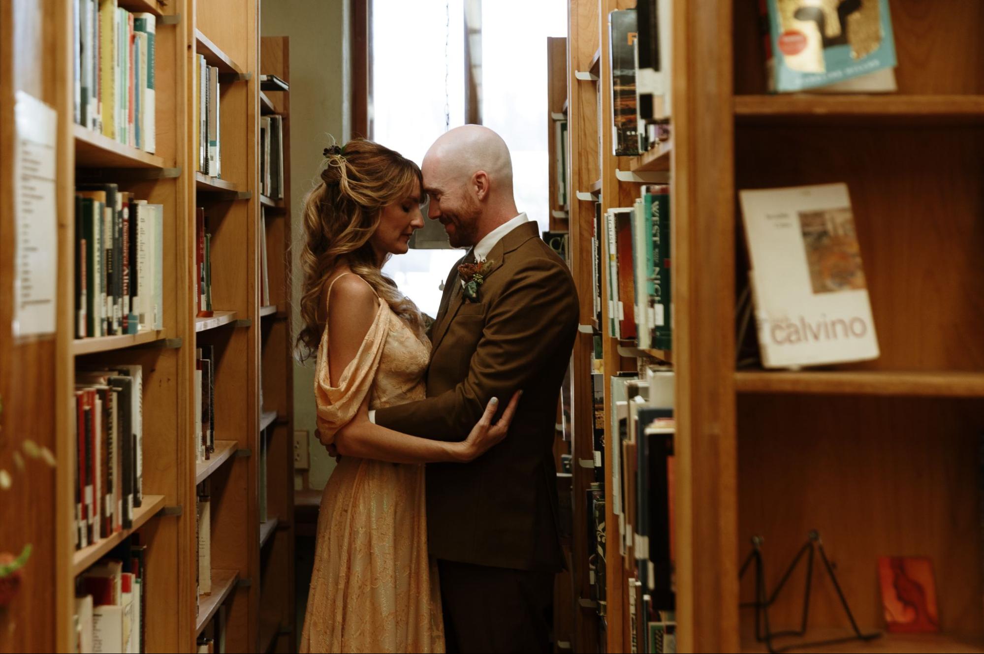 a bride and groom embracing between rows of bookshelves in a cozy library setting, showcasing a unique and personal way to personalize your wedding.