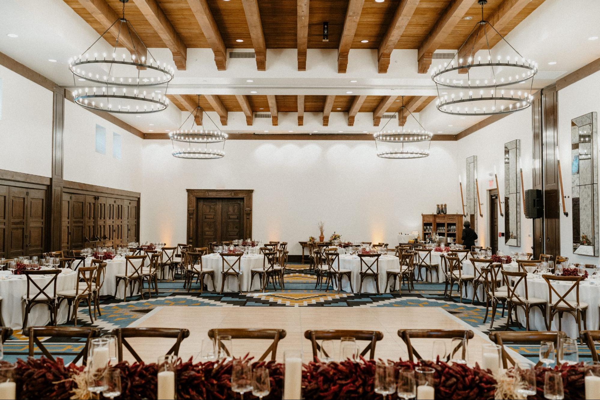 Elegant indoor reception area at one of the Santa Fe wedding venues, featuring a spacious room with wooden beams, large circular chandeliers, and round tables set with white linens. The decor includes rustic wooden chairs, candles, and floral arrangements.