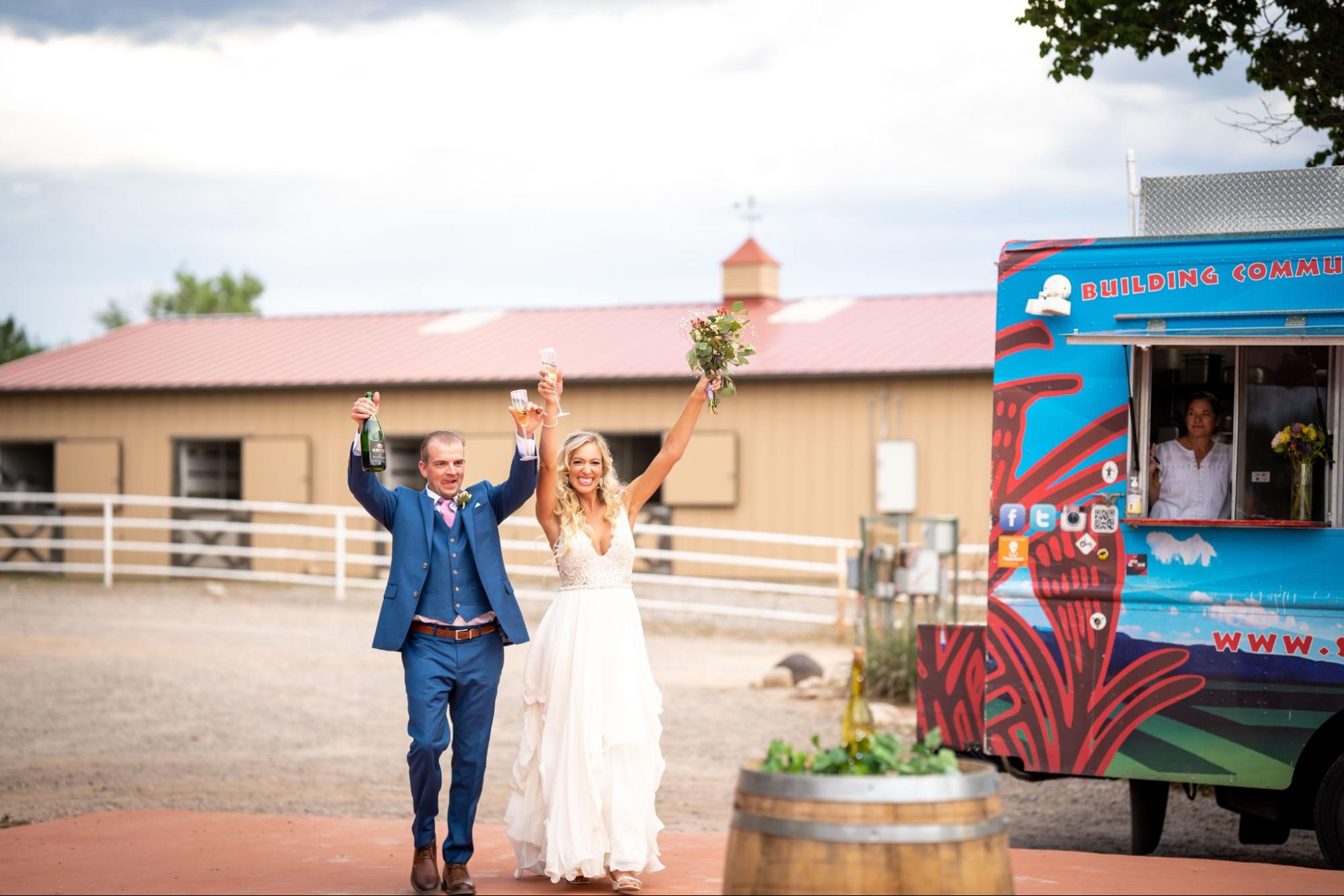 A joyful bride and groom celebrate with raised glasses, standing near a colorful food truck at an outdoor venue with rustic barn buildings in the background. A relaxed, festive way to make out-of-town guests feel at home at your Santa Fe wedding.