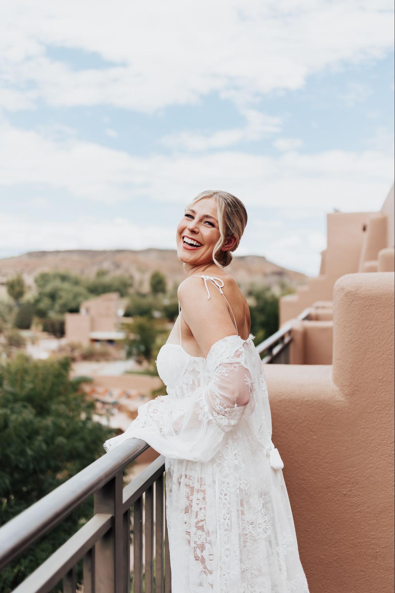 Bride laughing on a balcony with blue skies and adobe buildings in the background, wearing a lace off-the-shoulder gown. | outdoor wedding venues in albuquerque