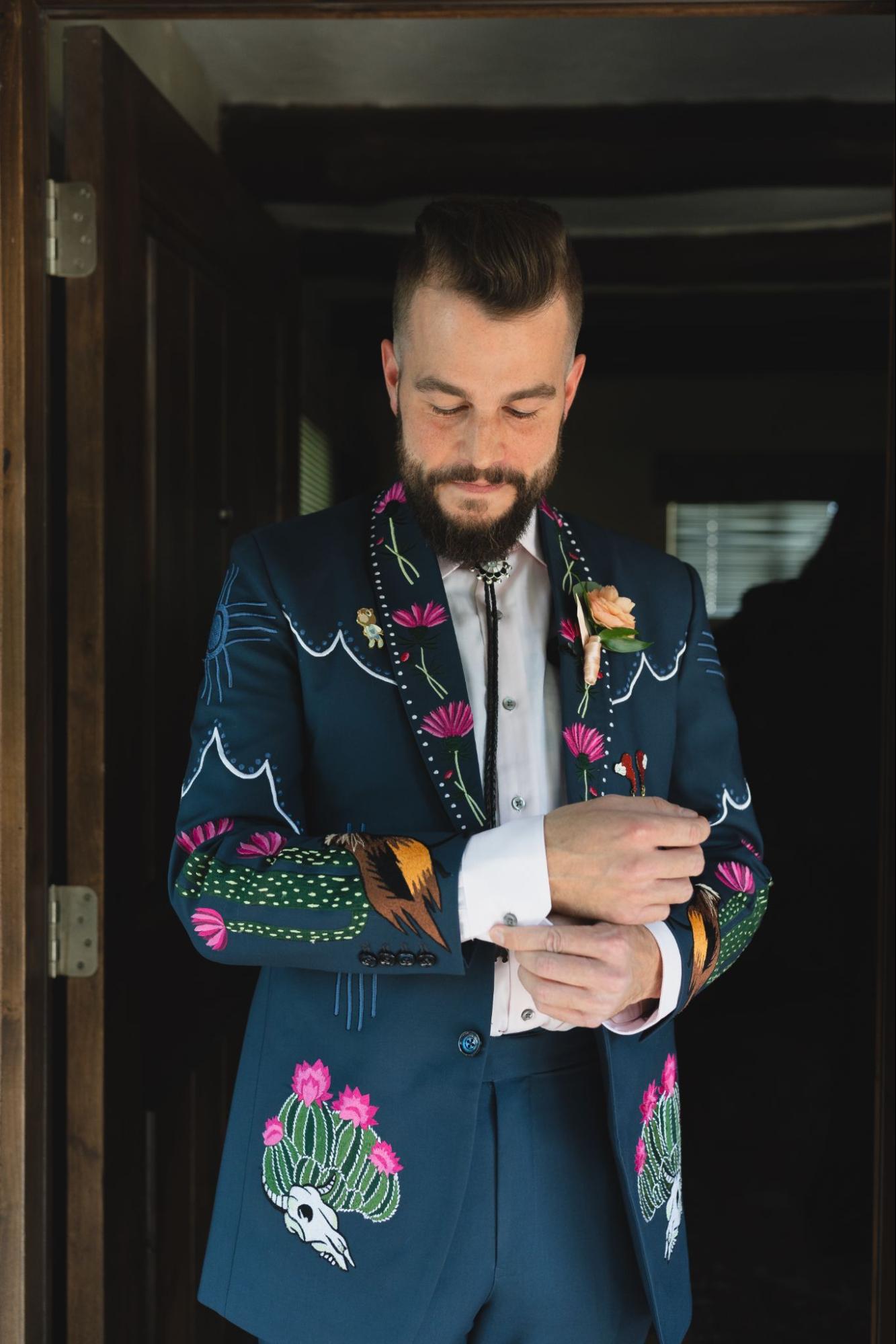 A groom adjusts his cufflinks while wearing a dark teal suit with colorful embroidery featuring cacti, flowers, and skull motifs.