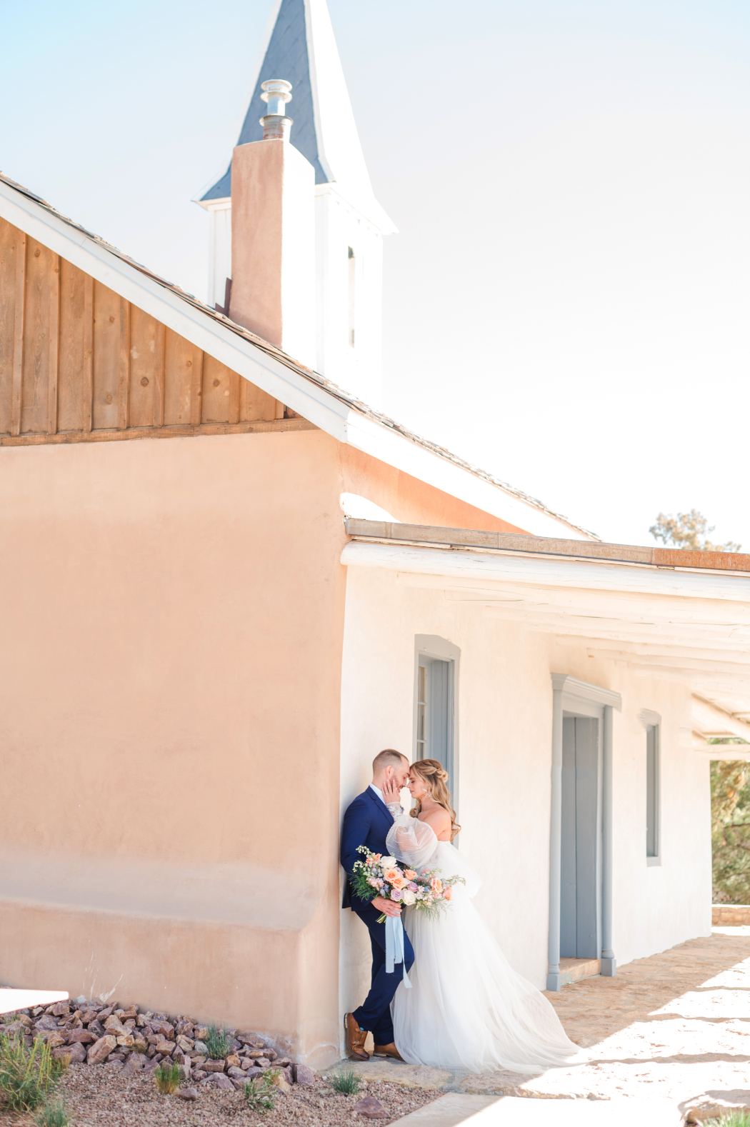 A bride and groom embrace beside the historic chapel at Bishop's Lodge Santa Fe, NM, with soft adobe tones and blue accents.