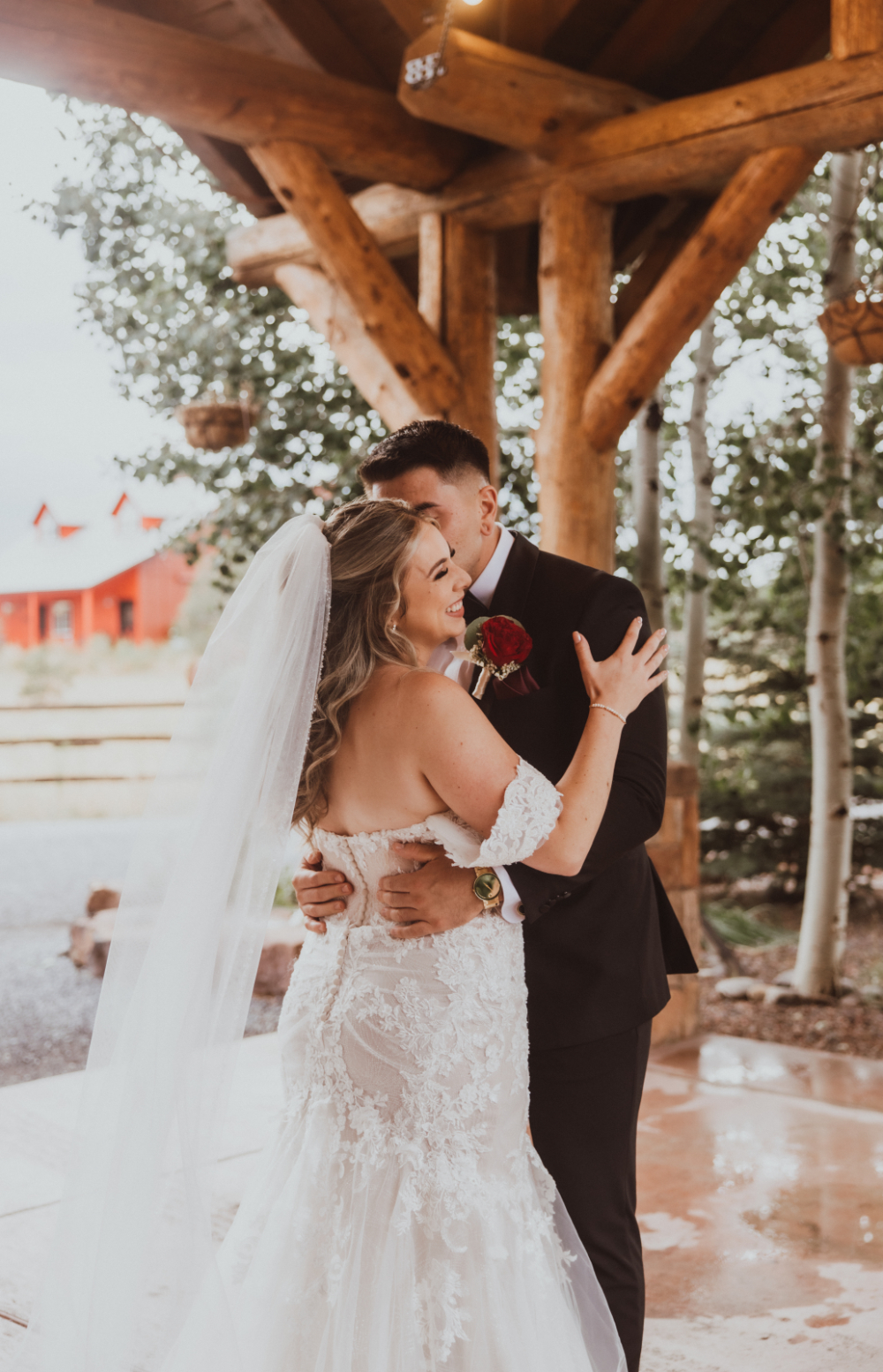 Bride and groom share a romantic embrace under a rustic wooden structure with greenery and a red barn in the background.