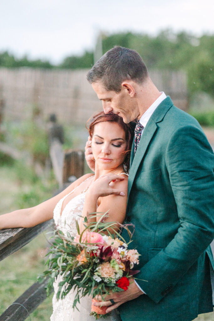 Groom lovingly leans his head on the bride's, as she holds a colorful bouquet by a wooden fence.