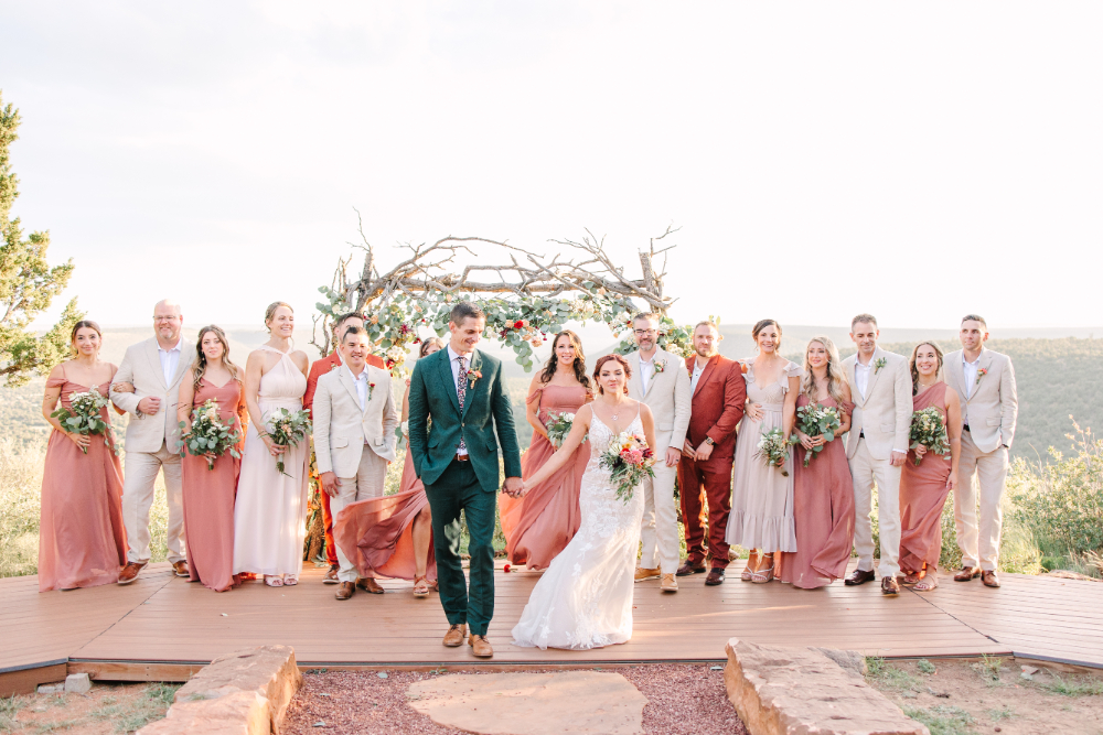Wedding party stands on an outdoor platform, with the couple leading at the center and a rustic arch decorated with greenery and flowers in the background | first steps after getting engaged