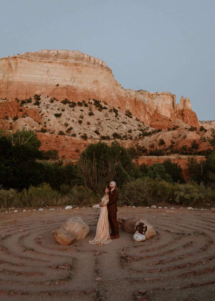 Couple embracing in a desert labyrinth at Ghost Ranch. The bride wears a boho gown, and the groom is in a dark suit, surrounded by red rock cliffs and desert greenery.