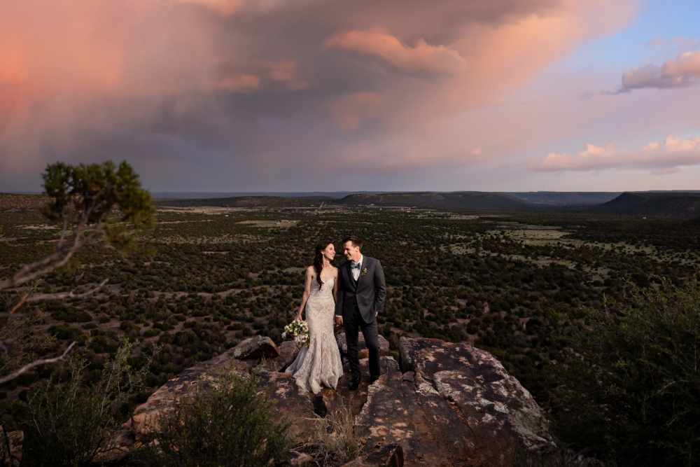 Bride and groom on a rocky overlook at sunset. The bride wears a lace gown and holds a bouquet, while the groom is in a dark suit, with vast desert views behind them.