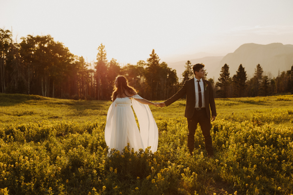 Couple walking through a wildflower meadow at golden hour. The bride wears a flowing white dress, and the groom is in a dark suit, with mountains in the background.