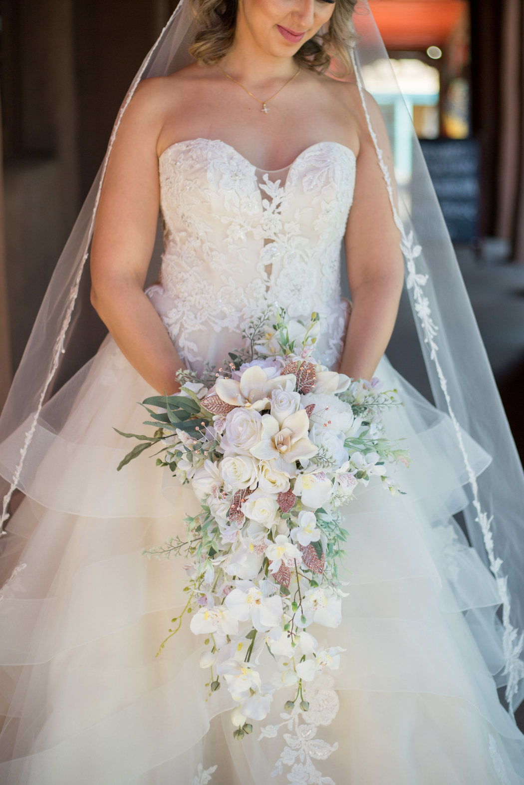 A bride in a strapless lace wedding dress holds a cascading white floral bouquet with roses and orchids, with a delicate lace-edged veil draped over her shoulders | best photographers in santa fe
