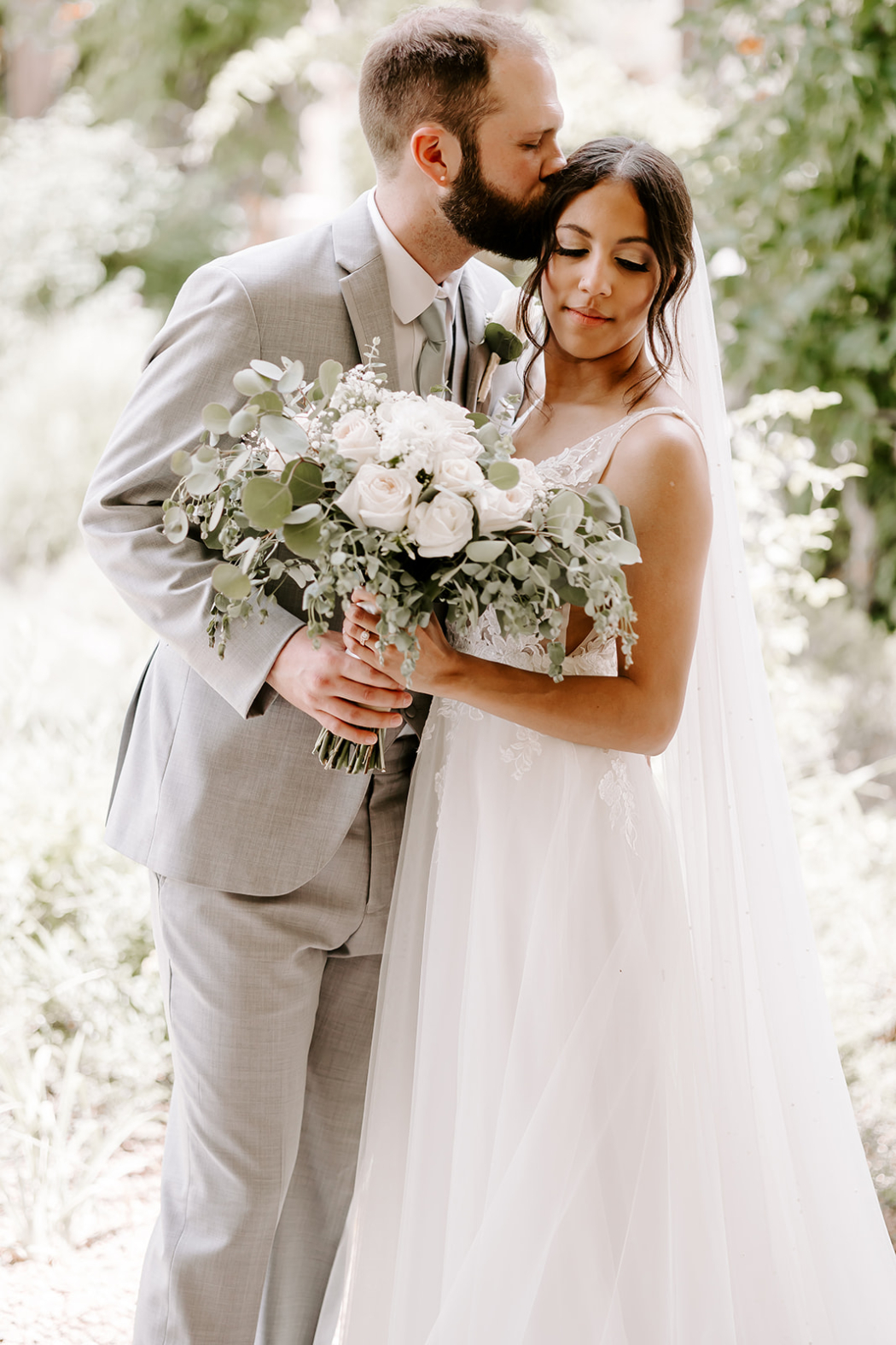 a bride and groom in an intimate pose, with the groom in a light gray suit kissing the bride’s forehead. She wears a flowing white gown with lace details and holds a bouquet of white roses and eucalyptus.