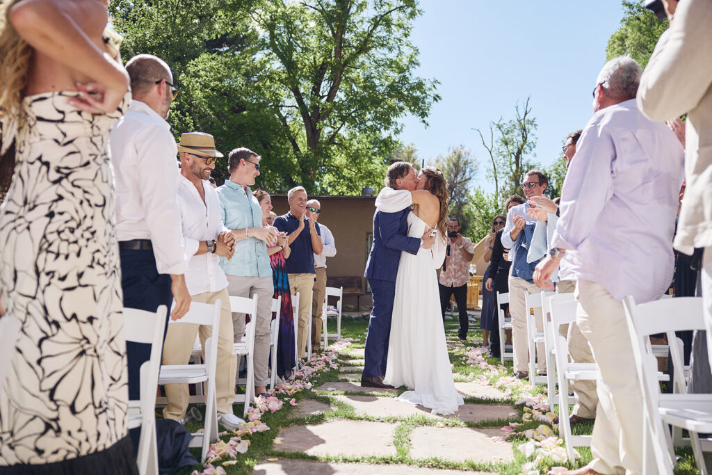 A newlywed couple shares a kiss at the end of the aisle as their guests cheer and clap around them.