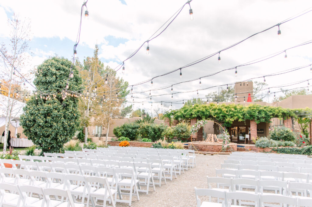 A full ceremony setup under string lights at La Posada in Santa Fe, featuring white chairs, lush greenery, and a floral arch backdrop.