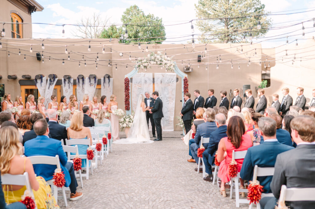 A courtyard ceremony at La Posada in Santa Fe, featuring adobe walls, string lights, and a floral arch backdrop.