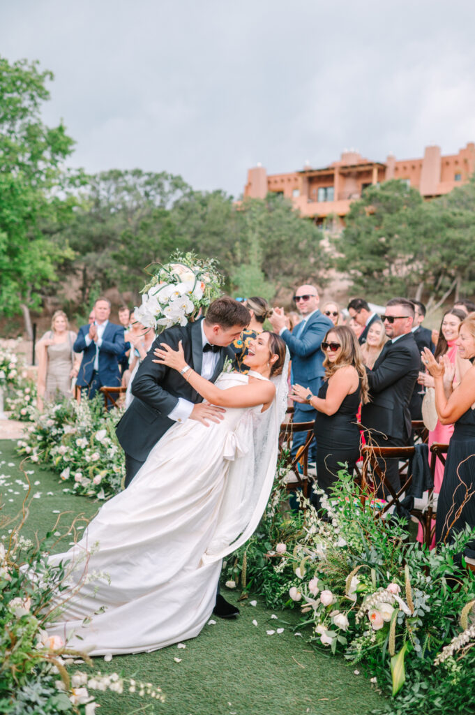 Groom dips bride as guests cheer during an outdoor ceremony.