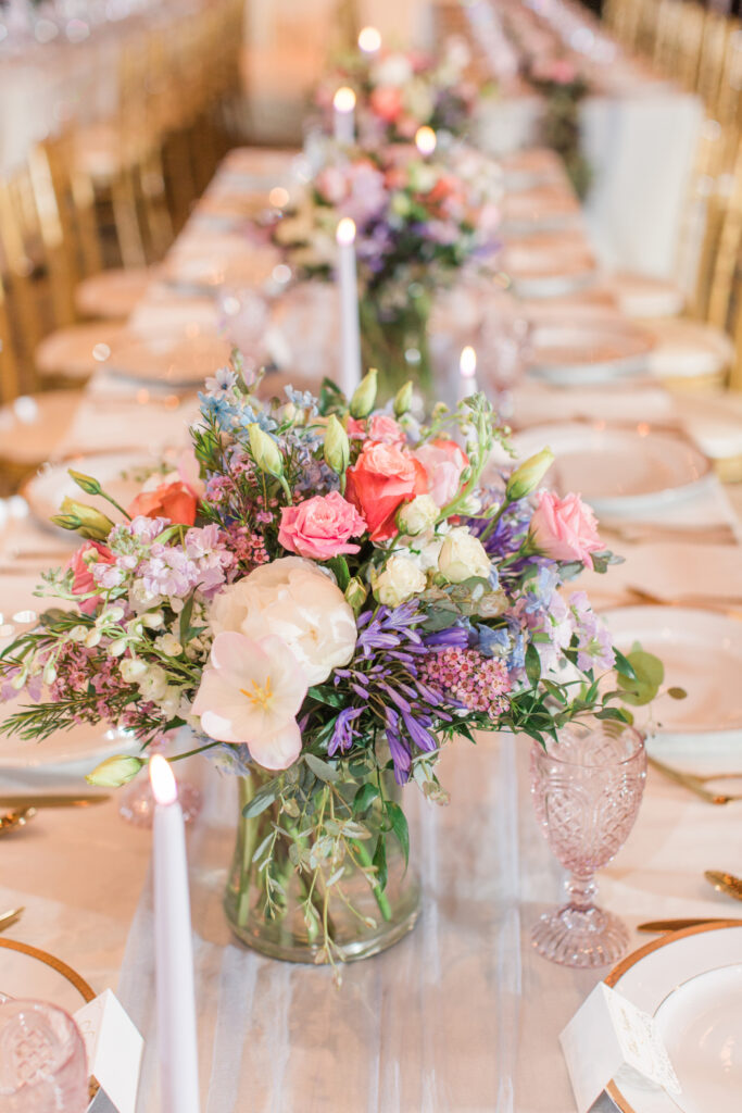 A soft spring centerpiece with blush, lavender, coral, and cream blooms sits in a glass vase on a long reception table, accented by tapered candles and gold-rimmed plates.