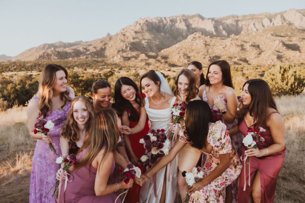 The bride and bridesmaids, in various shades of red, lavender, and floral prints, laugh together with the Sandia Mountains behind them—carrying simple bouquets of red and white blooms.