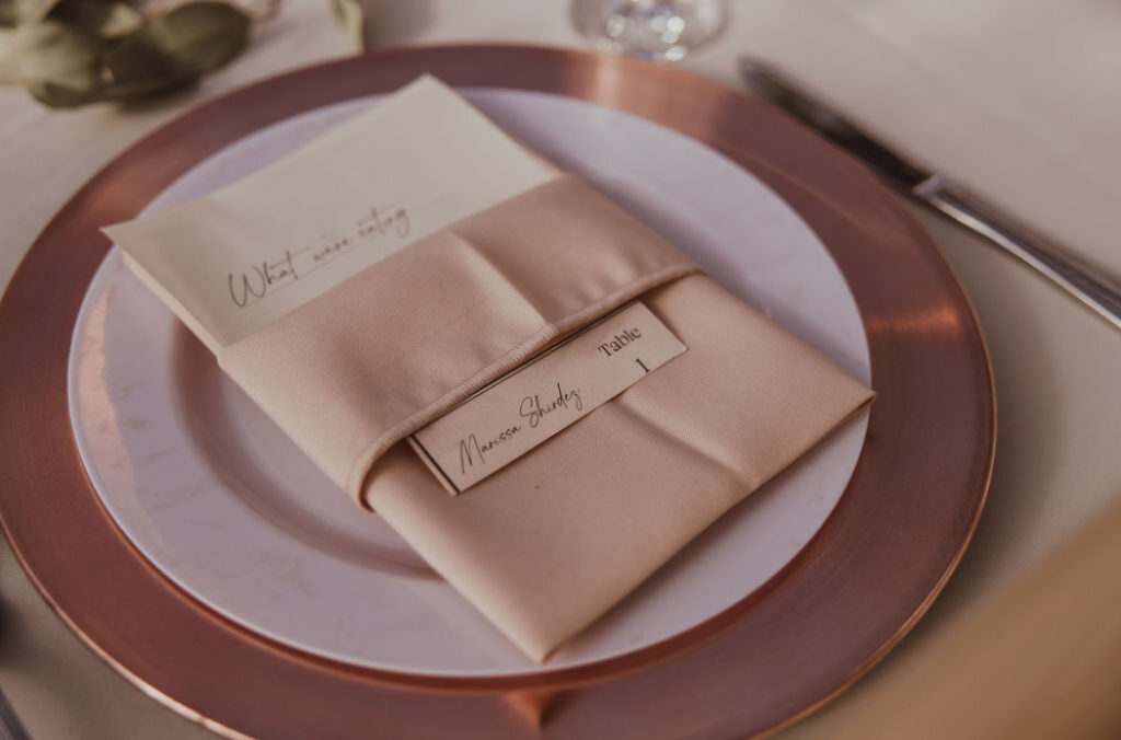 A minimalist place setting features a blush napkin folded around a name card and menu, sitting on a white plate with a copper charger. Simple and modern spring wedding color palettes.