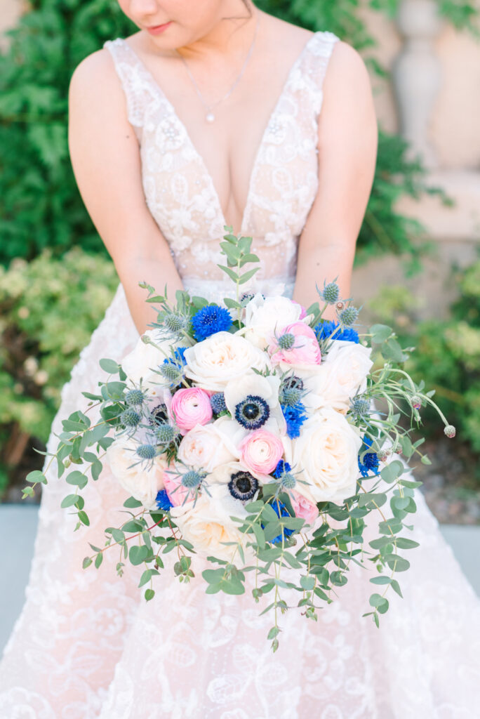 The bride holds a bouquet of white and blush roses, blue cornflowers, pink ranunculus, and greenery | Spring wedding color palettes.