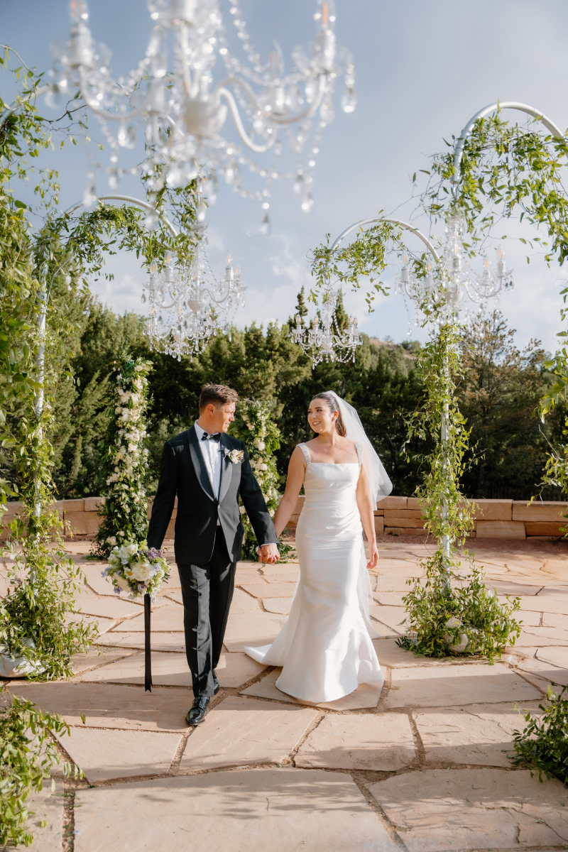 Bride and groom walking down aisle surrounded by lush greenery and chandelier wedding ceremony aisle decor.