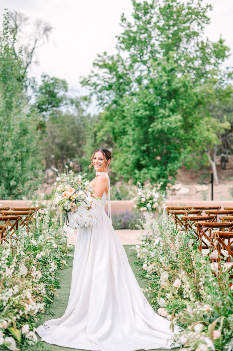 Bride stands in the center of ceremony aisle, holding luxury flower bouquet made for her Santa Fe celebration.