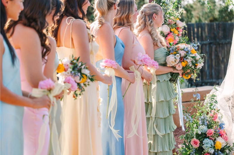 Bridesmaids in colorful dresses standing holding their bouquets during wedding ceremony.