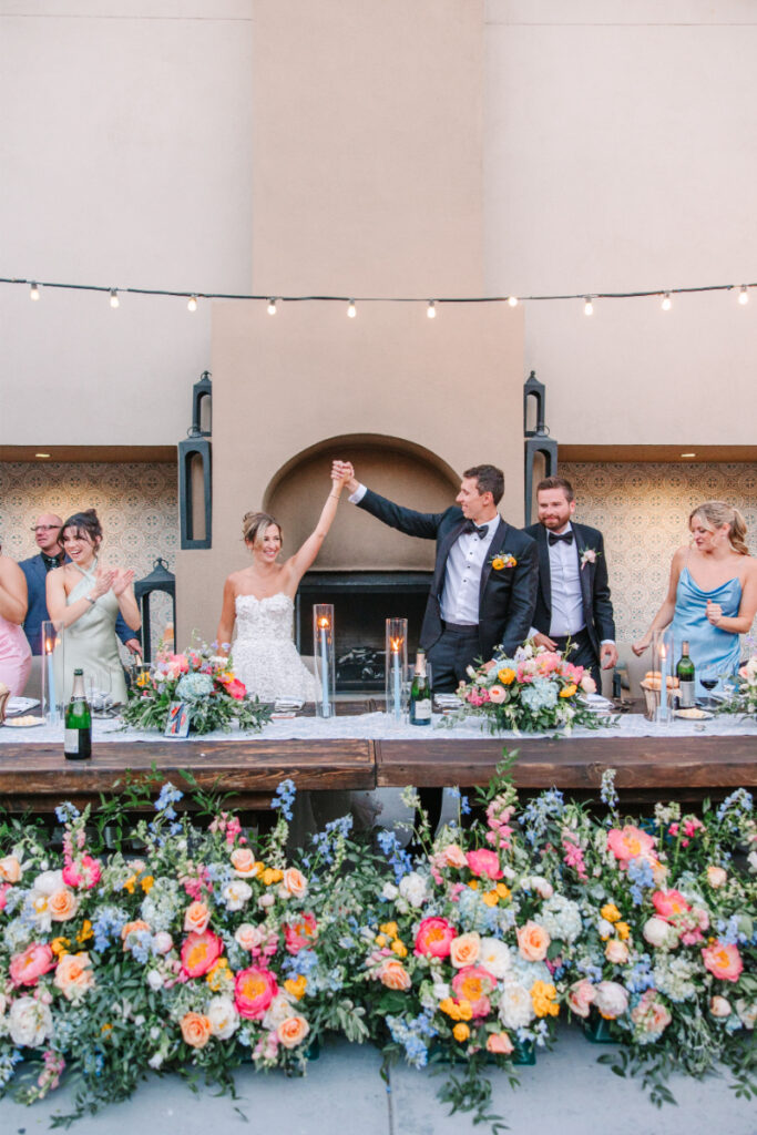 Bride and groom hold hands as they stand together in front of their wedding party table, surrounded by beautiful colorful florals arranged by one of the best Albuquerque florists.