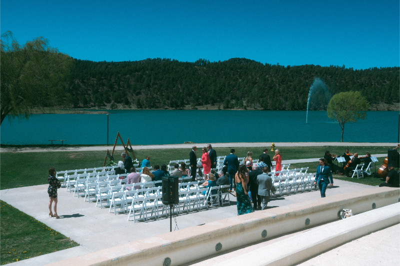 Guests get seated in white folding chairs for wedding ceremony on a lawn overlooking a lake.