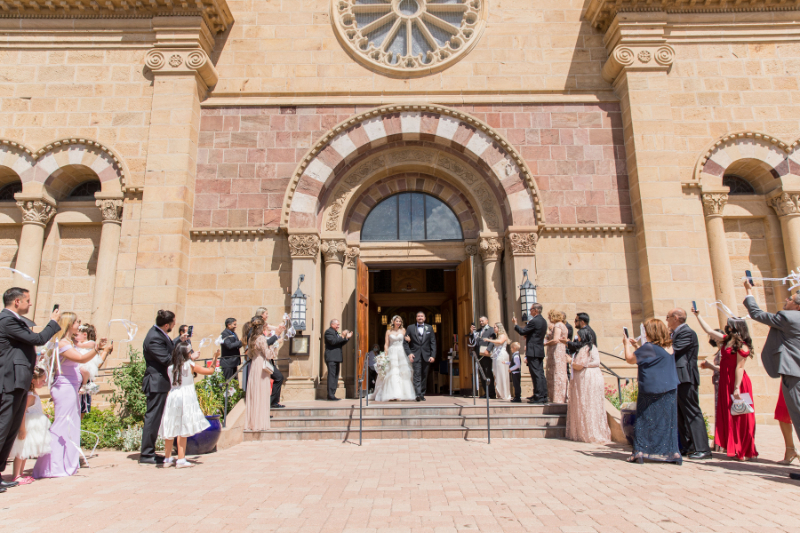 Bride and groom exit Loretto Chapel after their ceremony, guests excited.