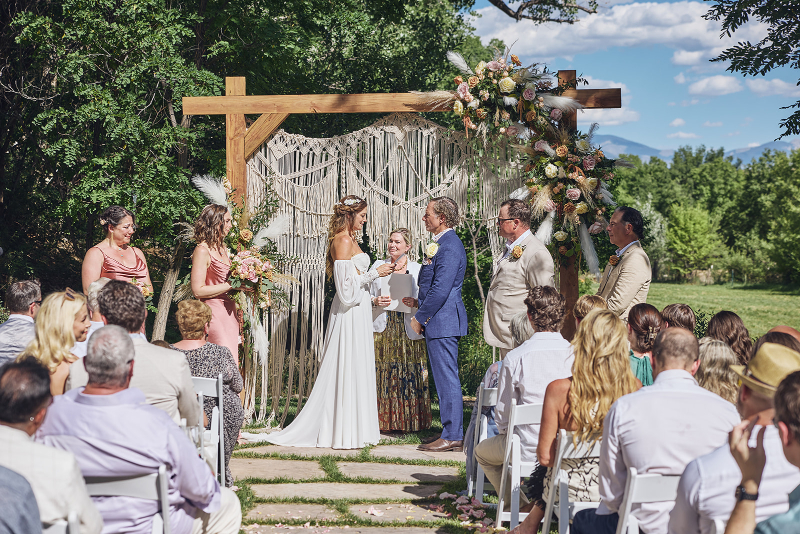 Bride and groom exchange vows in front of a wooden arch with macrame details, during their backyard wedding.