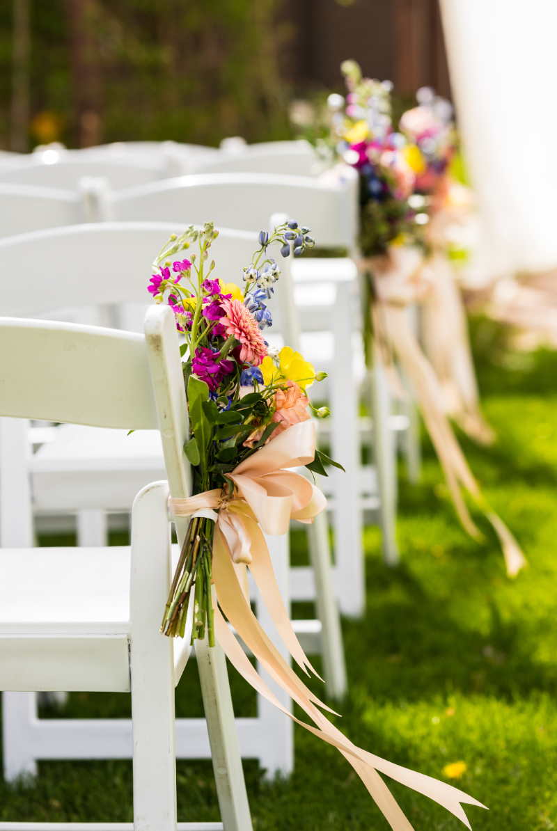 Colorful floral arrangement tied around white folding chair with salmon colored ribbon.