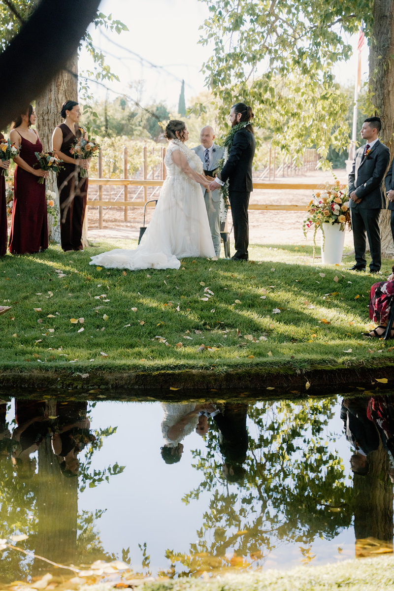 Bride and groom exchange vows on mansion wedding venues lawn with their reflection casted in the lake by them.