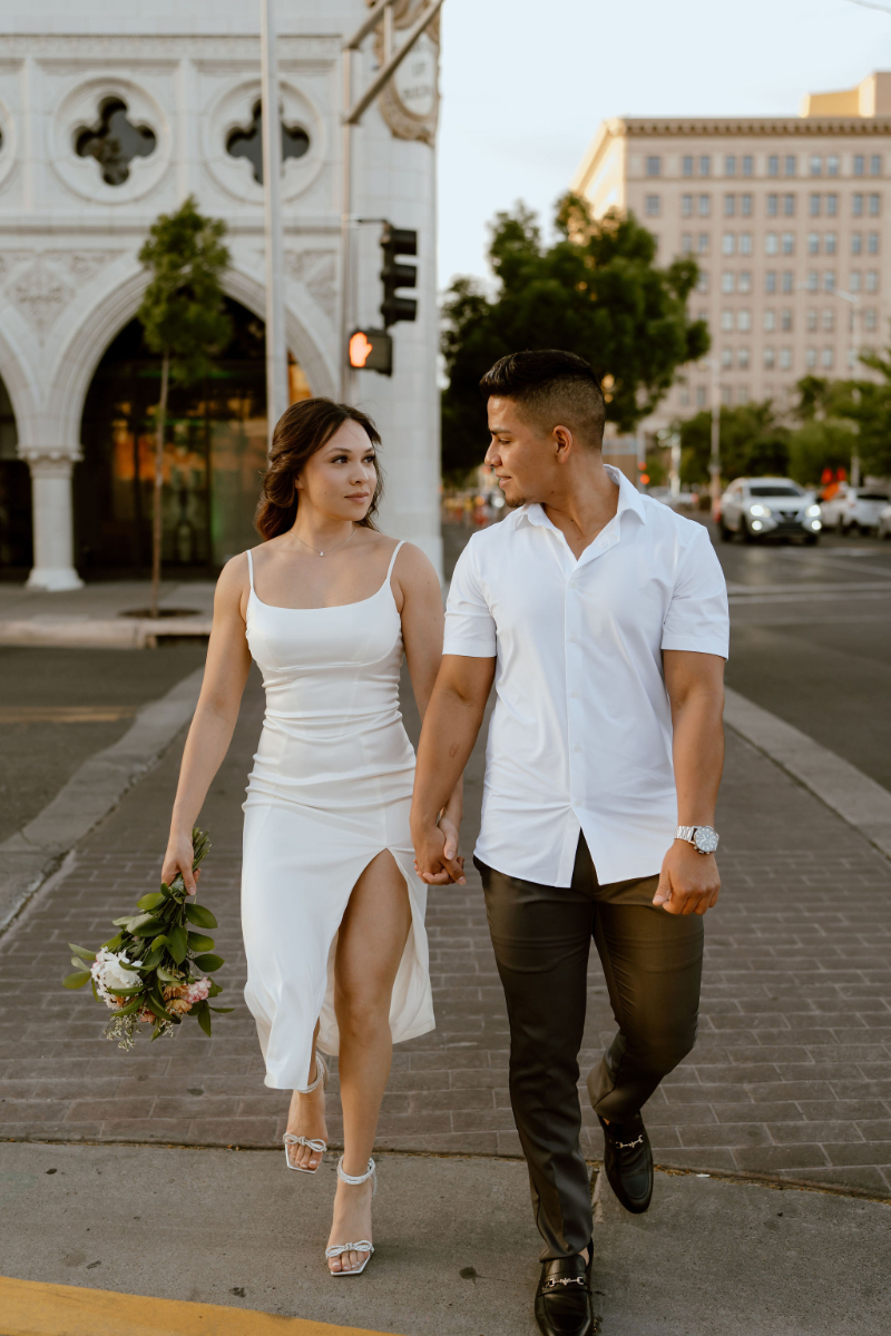 Couple looks at each other as they hold hands walking across the street during their engagement session in New Mexico.