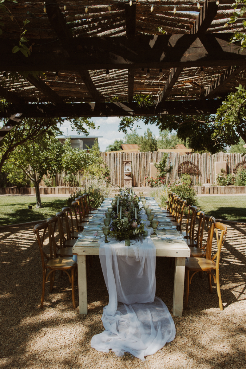 Long wooden table sits under pergola decorated with white table runner hanging to the ground, refined florals, and table settings for a French Chateau wedding style.