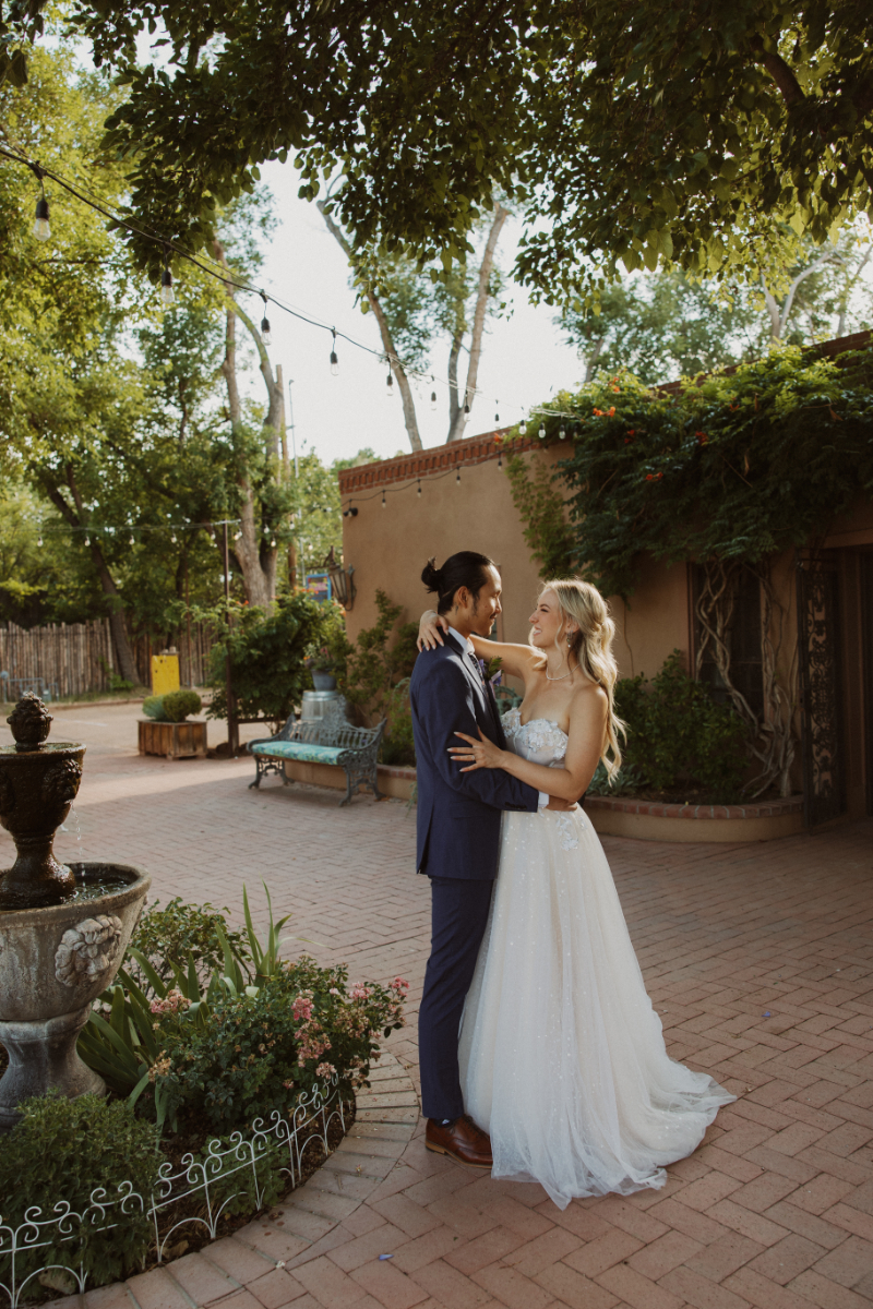 Bride and groom stand in a courtyard with their arms wrapped around each other as photographer captures their wedding portraits.