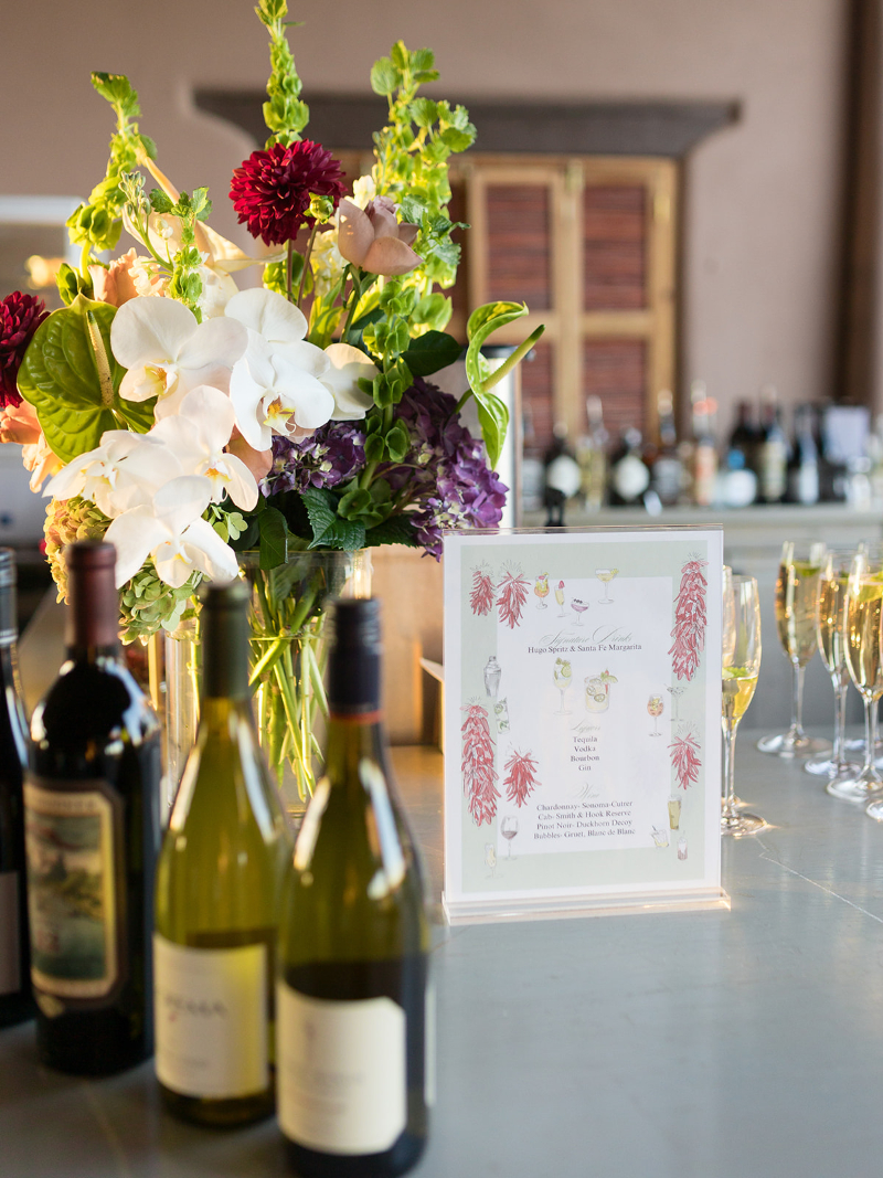 Santa Fe style bar menu for wedding sitting next to floral arrangement and wine bottles.