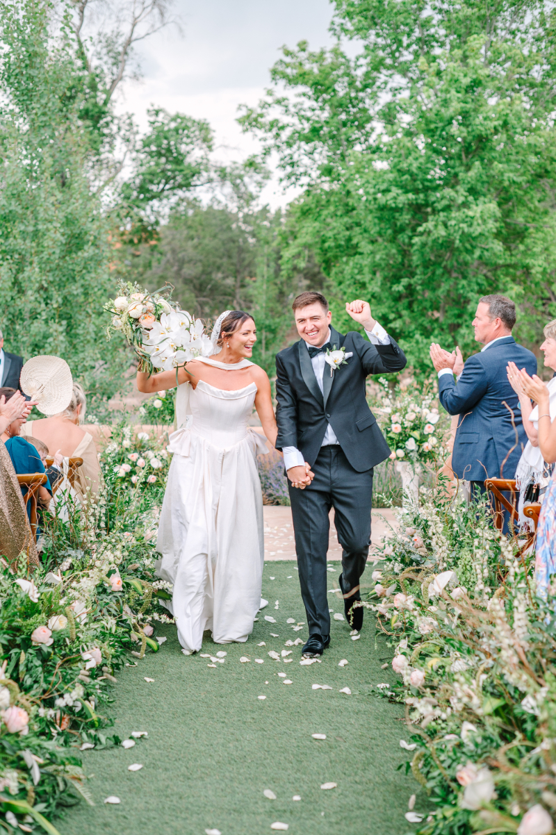 Bride lifts her bouquet in the air as she processes with her husband down the aisle.