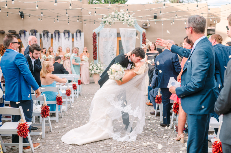 Groom dips bride for a kiss and they recess down aisle, guests cheering.