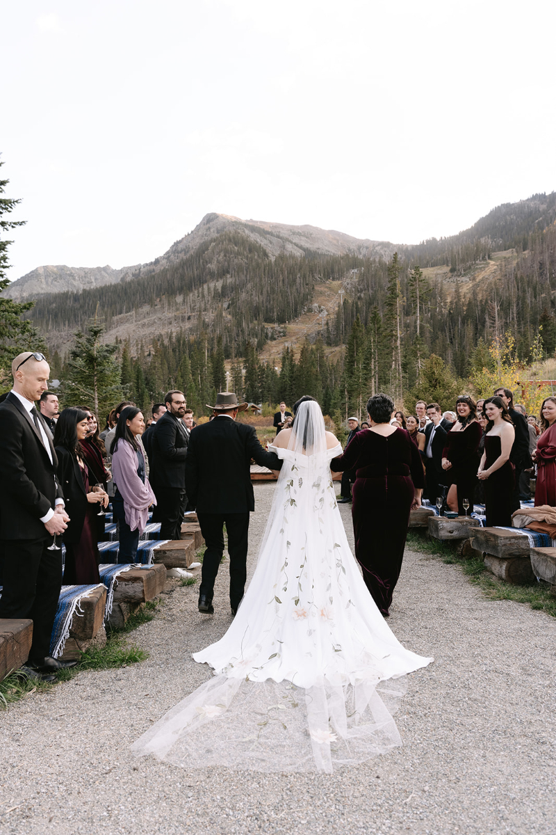 Bride is walked down the aisle by both parents, her floral embroidered veil dragging behind her as she walks faces the mountains at her Knoxville wedding venues.