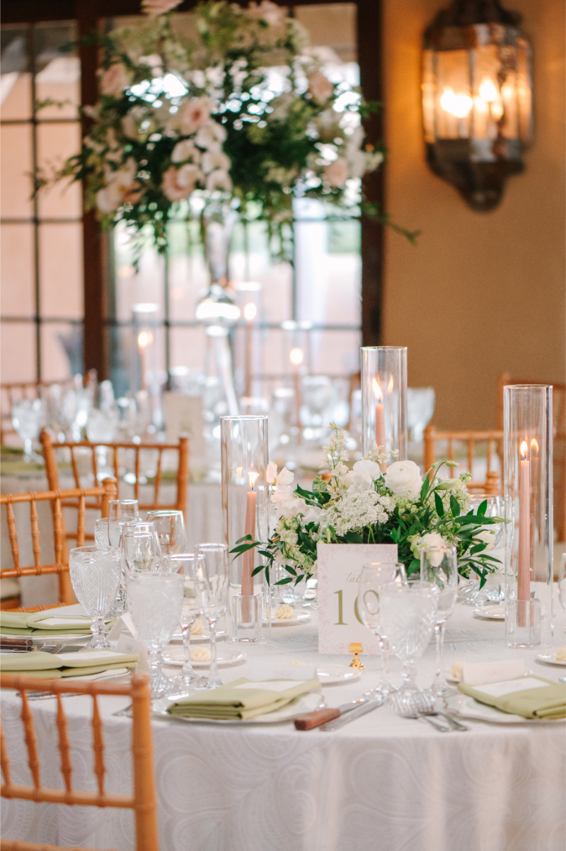Tablescape decorated with spring centerpiece ideas including white floral arrangement and pillar candles in hurricanes.
