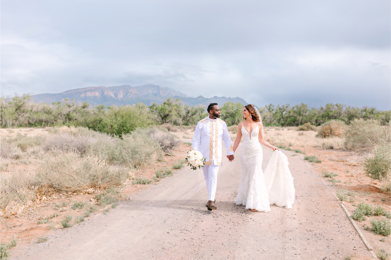 Bride and groom walk hand in hand through the desert, the sky full of clouds. They answered the question of "what time do weddings start" well when they planned their wedding in New Mexico.