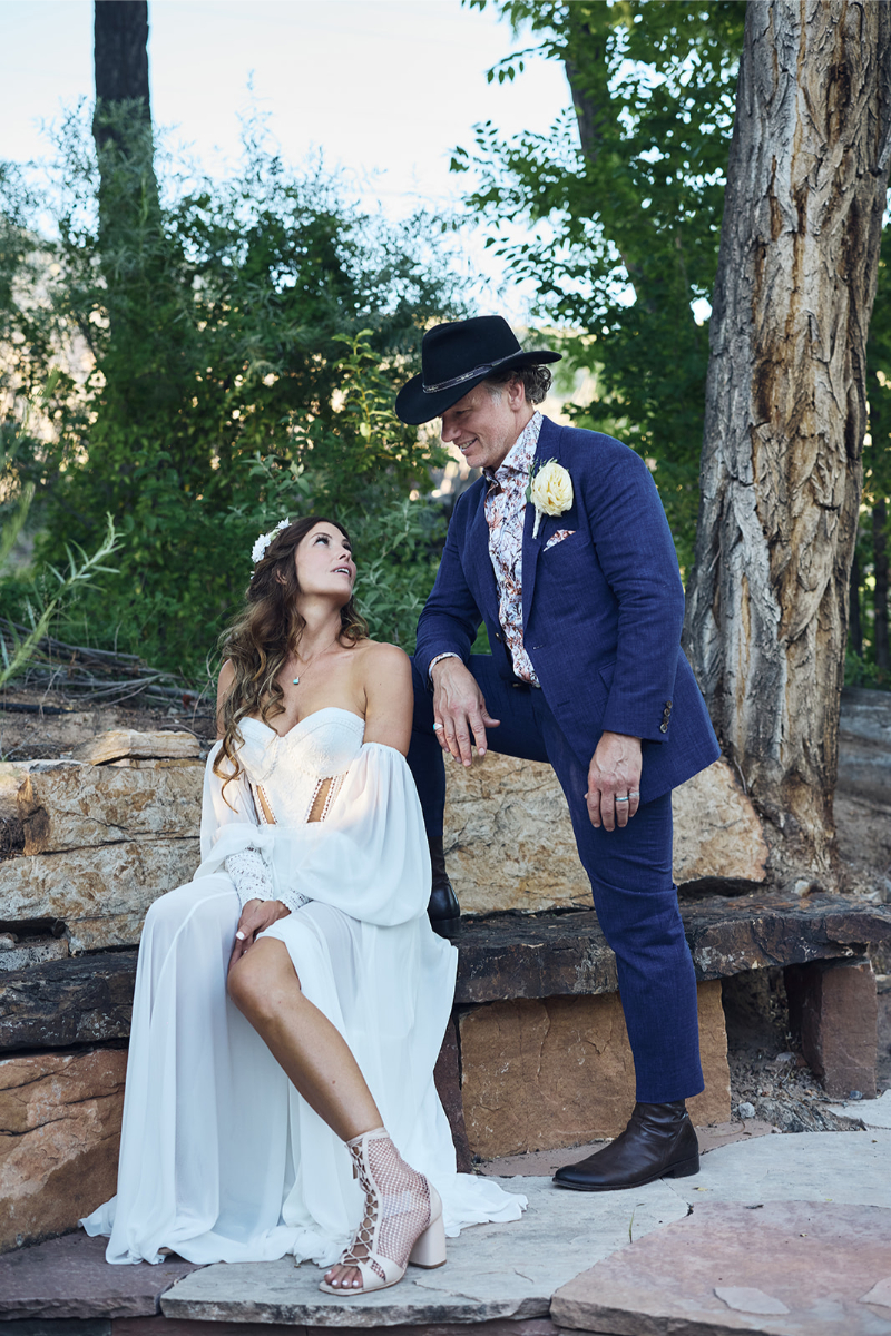 Bride sits on rock and looks up at groom who set his foot on the bench and is resting his hand on his knee.