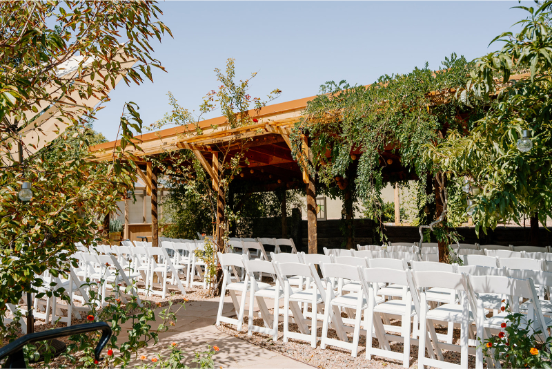 Desert wedding ceremony in New Mexico set up under vine-covered awning and white folding chairs.