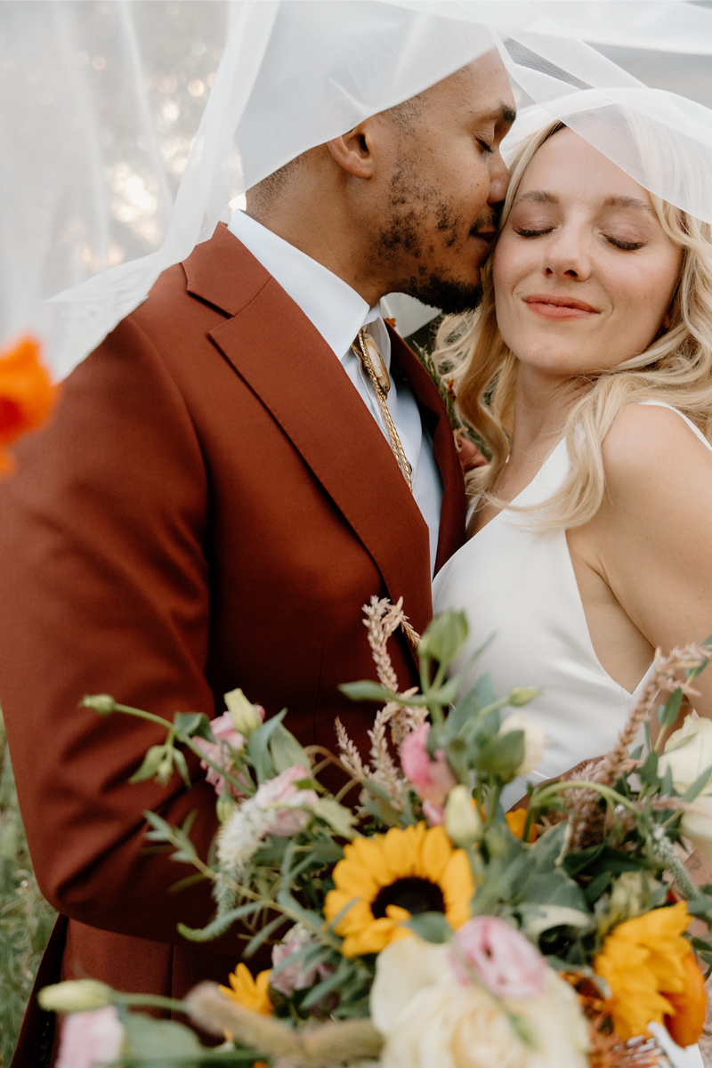Groom kisses bride on the top of her head as they take portraits under her veil after their desert wedding ceremony in New Mexico.