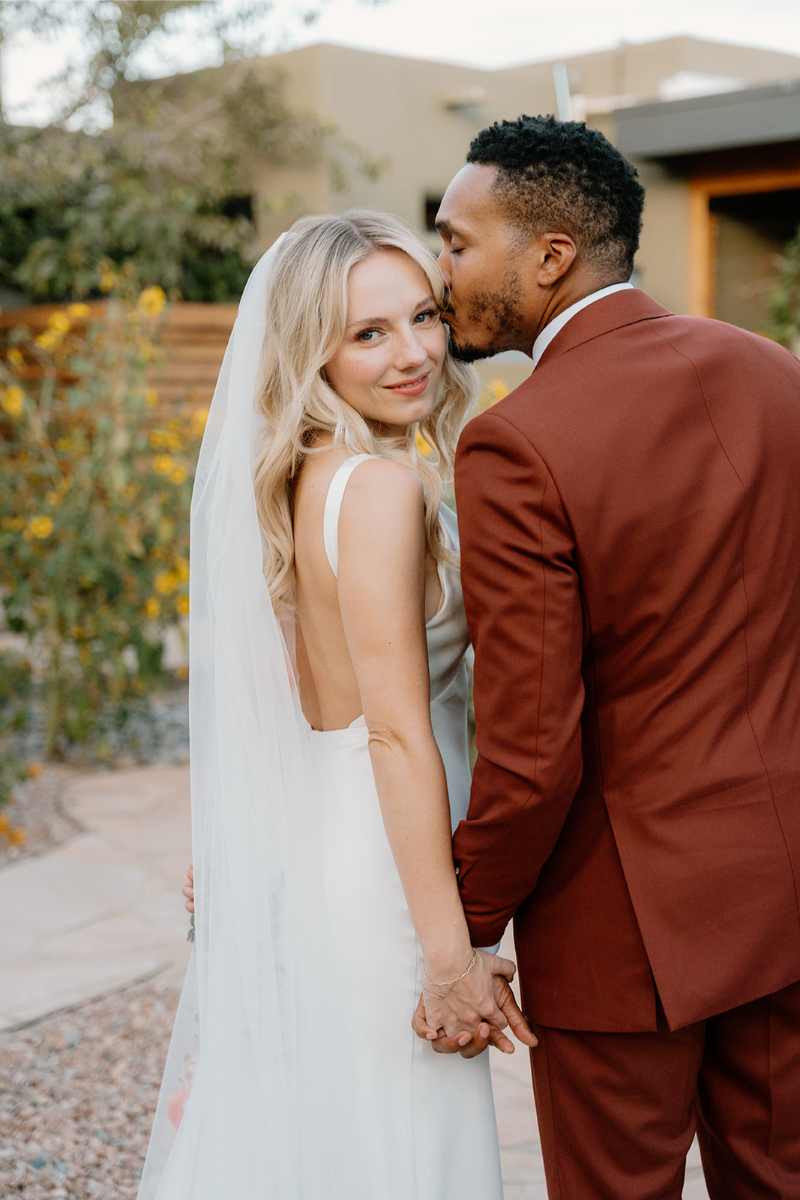 Groom kisses bride on the top of her head as they take portraits after their desert wedding ceremony in New Mexico.