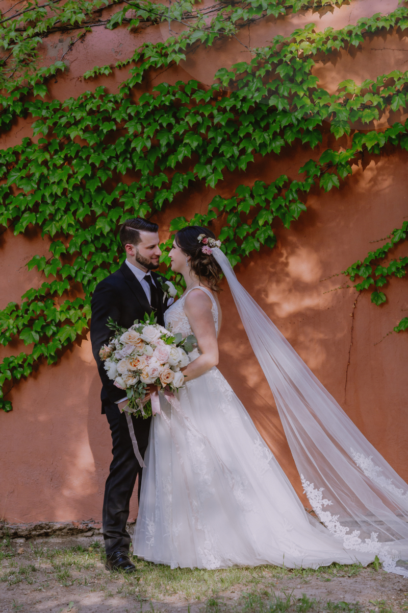 Bride and groom stand for wedding portraits in front of terracotta colored wall covered in vines.
