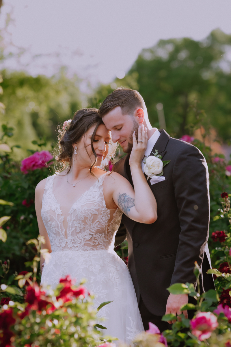 Bride stands in front of groom, their foreheads touching, as she lifts her hand to his cheek, standing in front of pink rose bushes.