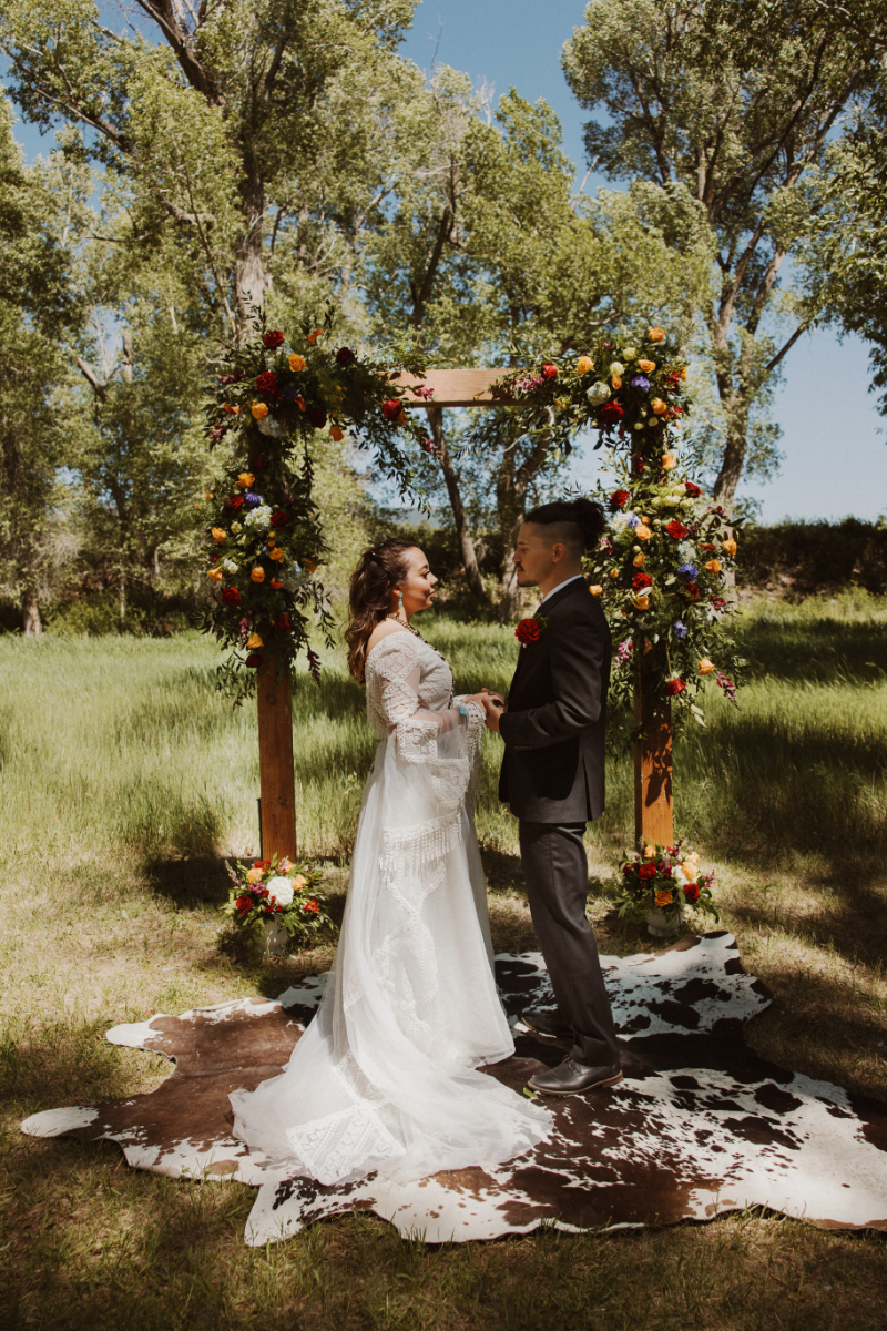 Bride and groom exchange vows while standing on a brown cow hide rug, surrounded by fall florals on a rectangular arbor.